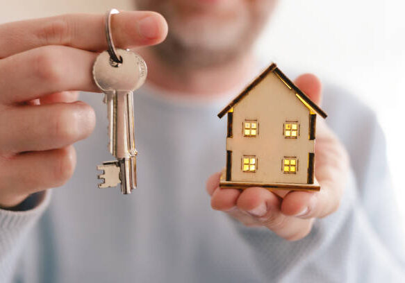 Close up color image depicting a new home owner (a man in his mid to late 30s, Caucasian ethnicity) holding a set of house keys in one hand, and a wooden model house in the other. Room for copy space.