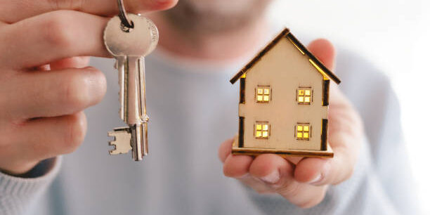 Close up color image depicting a new home owner (a man in his mid to late 30s, Caucasian ethnicity) holding a set of house keys in one hand, and a wooden model house in the other. Room for copy space.