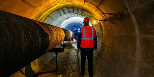 Tunnel worker examines pipeline in underground tunnel.